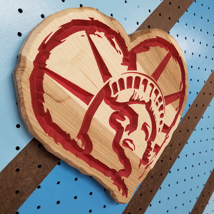 Angled shot of a relief carved hardwood heart-shaped sign featuring a detailed Statue of Liberty profile design painted red.