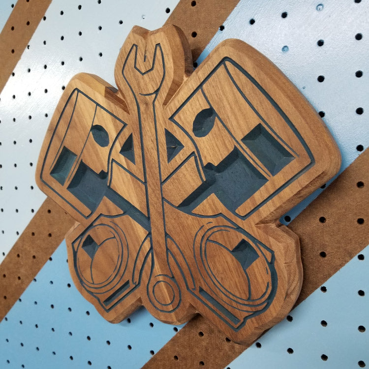 Angled shot of a relief carved hardwood sign with crossed pistons and a wrench design on a striped pegboard.