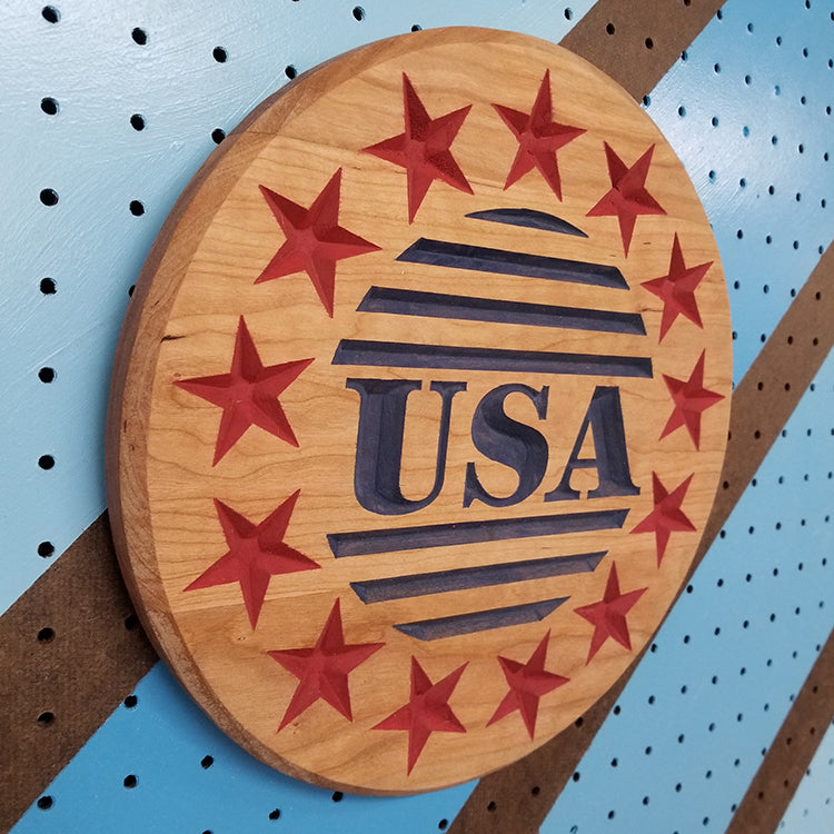 Angled shot of a custom hardwood USA Circle patriotic sign with a carved ring of stars and center relief text on a blue pegboard.