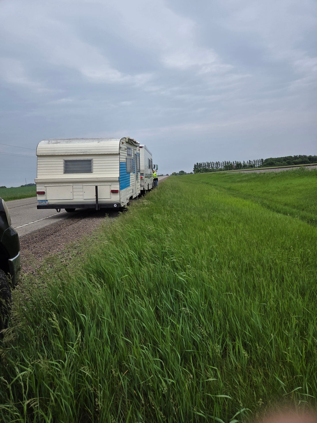 1981 Winnebago hauling the 1963 TravelEZE camper boutique for Whiskey River Craftworks along a rural Midwest roadside under cloudy skies.