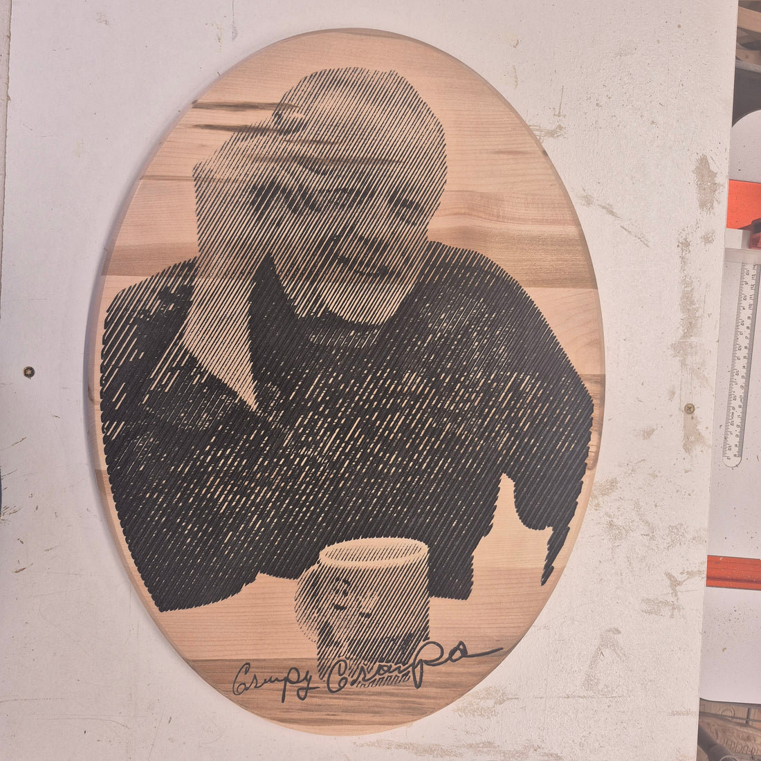 A detailed CNC-carved wooden oval plaque featuring a custom engraved portrait of an elderly man wearing a sweater and leaning his head on his hand next to a coffee mug. The natural wood grain shows through the detailed line work.