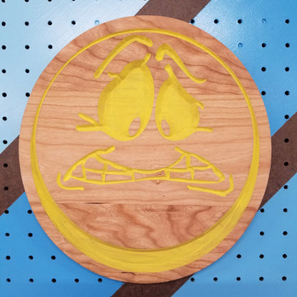 Custom hardwood worried face emoji sign with yellow painted relief carving on a blue shop pegboard.