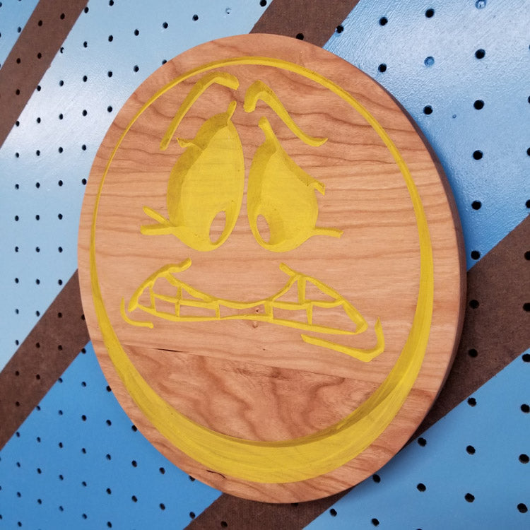 Angled shot of a custom hardwood worried face emoji sign with yellow painted relief carving on a blue pegboard.