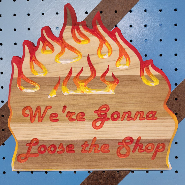 Relief carved hardwood sign with painted flames and We're Gonna Loose the Shop text on a blue and brown striped pegboard.
