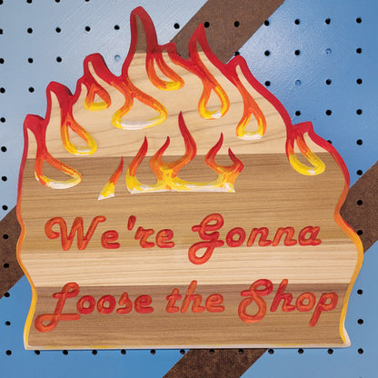 Relief carved hardwood sign with painted flames and We're Gonna Loose the Shop text on a blue and brown striped pegboard.