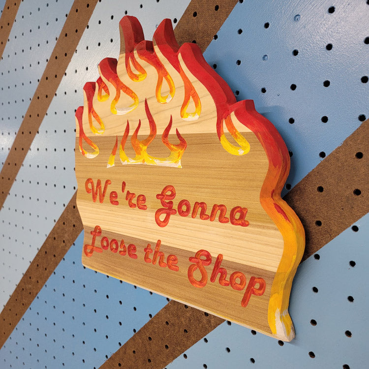 Angled shot of a relief carved hardwood sign with painted flames and We're Gonna Loose the Shop text on a blue and brown striped pegboard.