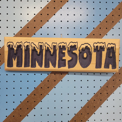 Relief carved hardwood sign with snow-capped MINNESOTA COLD text on a blue and brown striped pegboard background.
