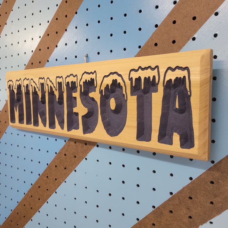 Angled shot of a relief carved hardwood sign with snow-capped MINNESOTA text on a blue and brown striped pegboard.