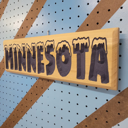 Angled shot of a relief carved hardwood sign with snow-capped MINNESOTA text on a blue and brown striped pegboard.