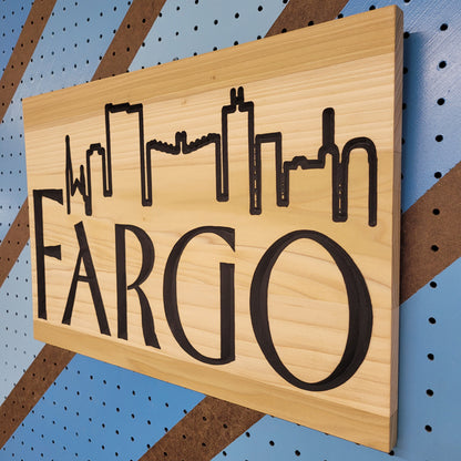 Angled shot of a relief carved hardwood sign featuring the iconic Fargo-Moorhead skyline silhouettes and Fraser-inspired lettering.