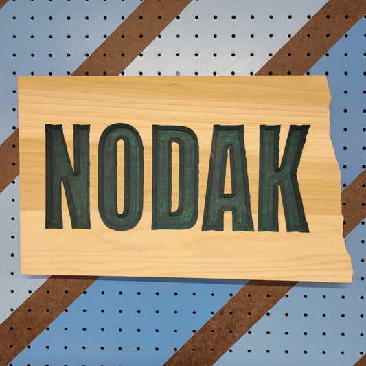 Relief carved hardwood sign in the shape of North Dakota with NODAK text on a blue and brown striped pegboard background.
