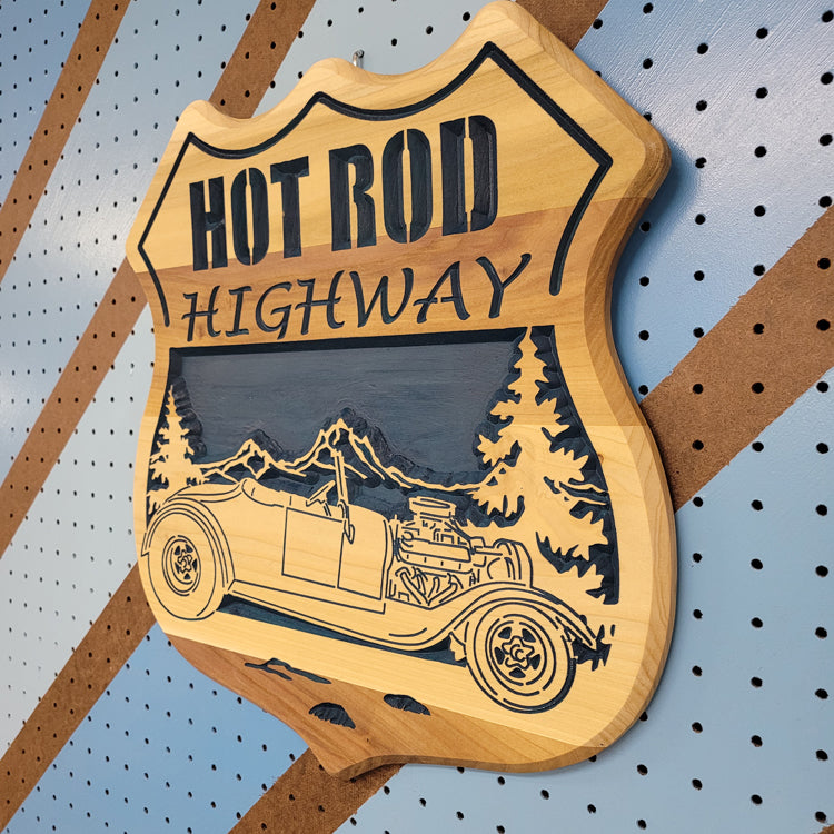 Angled shot of a fully customizable relief carved hardwood shield-shaped sign featuring a vintage hot rod convertible and pine trees.