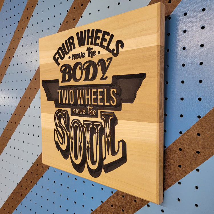 Angled shot of a Handcrafted relief-carved hardwood sign featuring the text "Four Wheels Move The Body, Two Wheels Move The Soul."
