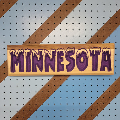 Relief carved hardwood sign with snow-capped MINNESOTA COLD text on a blue and brown striped pegboard background.