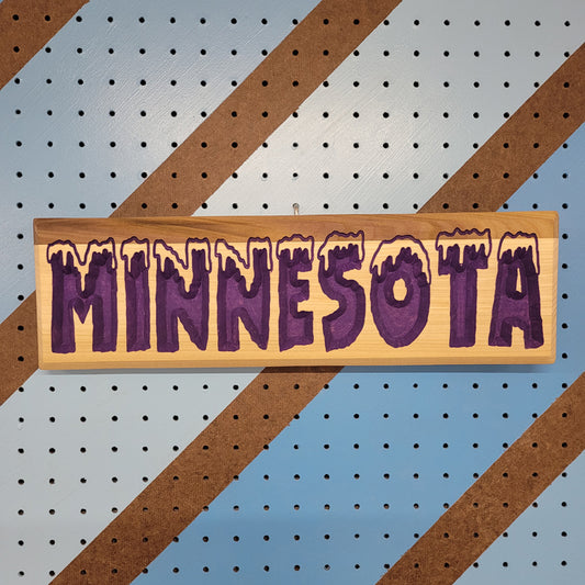 Relief carved hardwood sign with snow-capped MINNESOTA COLD text on a blue and brown striped pegboard background.