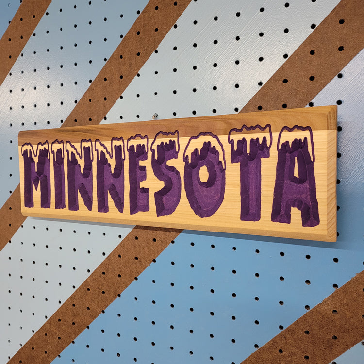 Angled shot of a relief carved hardwood sign with snow-capped MINNESOTA text on a blue and brown striped pegboard.