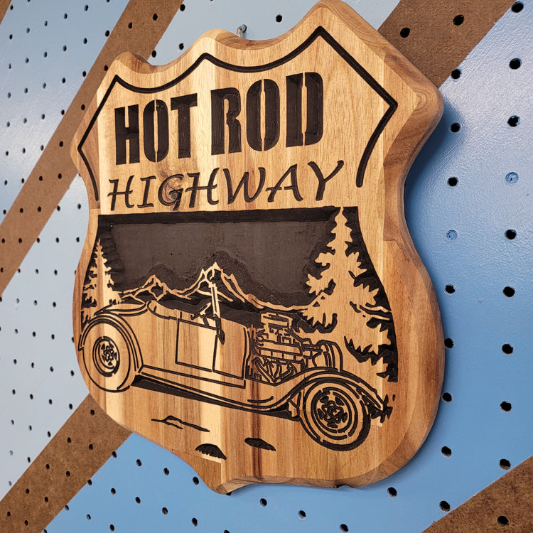 Angled shot of a fully customizable relief carved hardwood shield-shaped sign featuring a vintage hot rod convertible and pine trees.