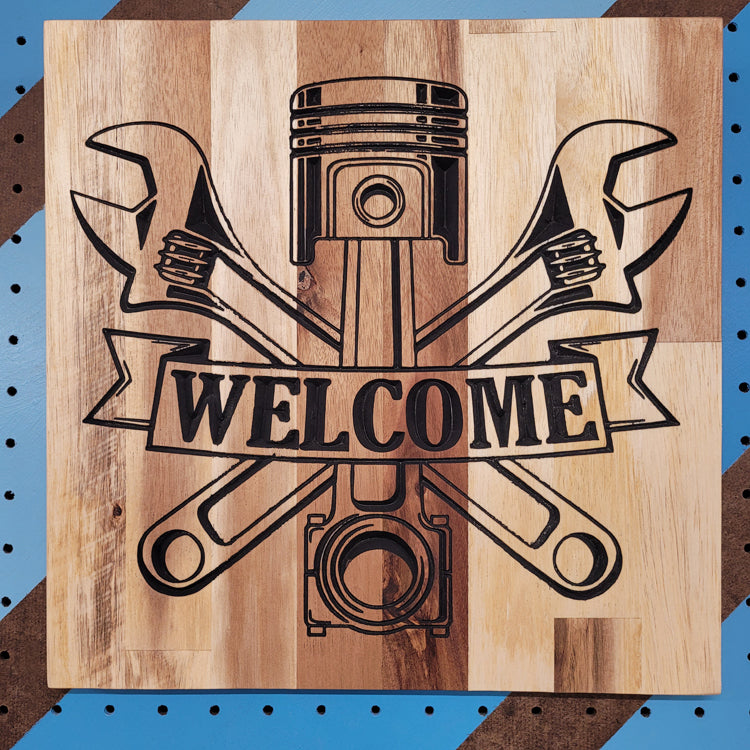 Custom hardwood Welcome garage sign with piston and crossed wrenches relief carving on a blue shop pegboard.