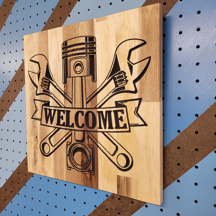 Angled shot of a custom hardwood Welcome sign with piston and crossed wrenches relief carving on a blue pegboard.