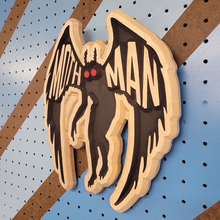 Angled shot of a relief carved hardwood sign with a detailed Mothman cryptid design on a blue and brown striped pegboard.