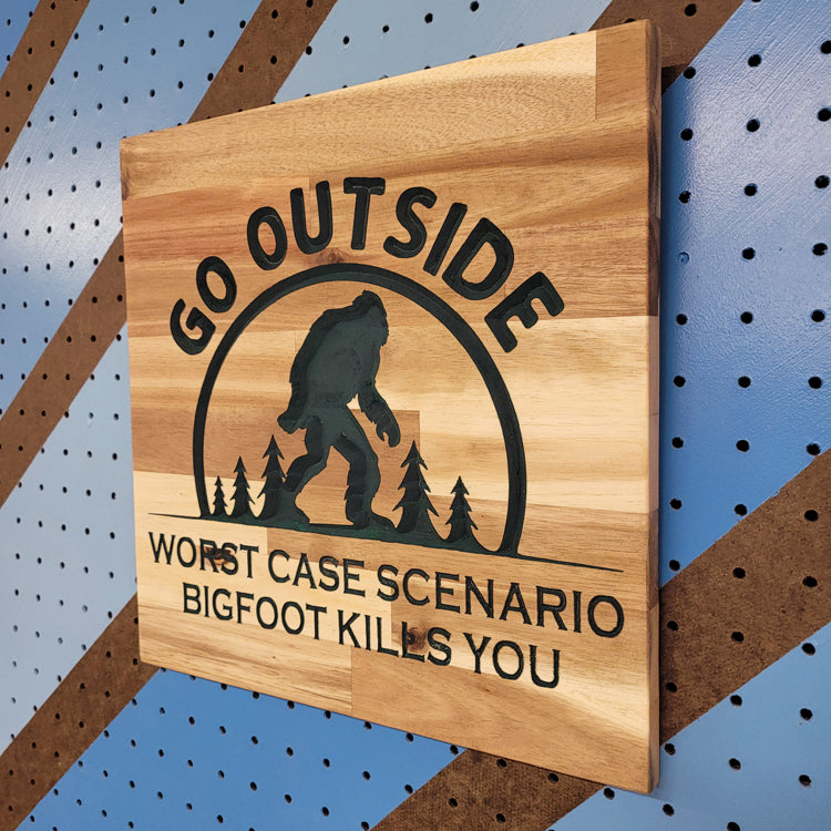 Angled shot of a Handcrafted relief-carved hardwood sign featuring a Bigfoot silhouette and the text "Go Outside, Worst Case Scenario Bigfoot Kills You."