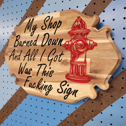 Angled shot of a custom hardwood sign with relief carved red fire hydrant and My Shop Burned Down text on a striped pegboard.