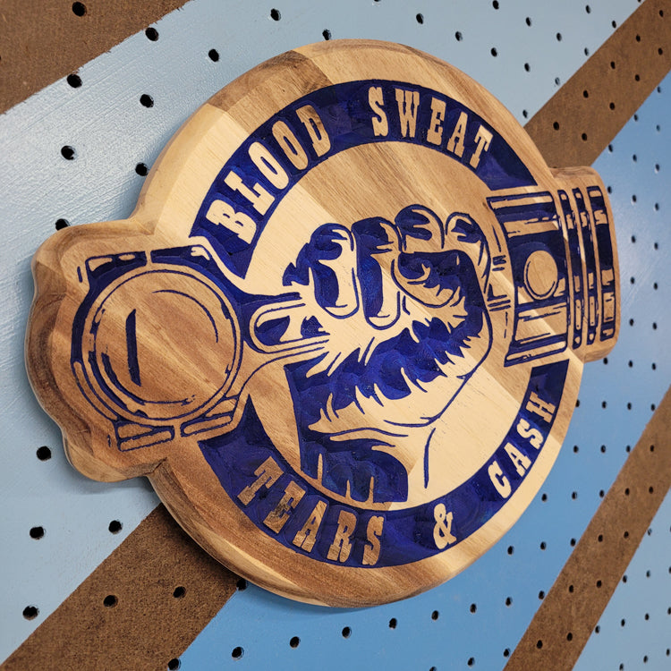 Angled shot of a relief carved hardwood sign with blue painted piston fist design and Blood Sweat Tears & Cash text on pegboard.