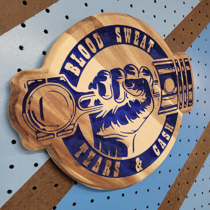 Angled shot of a relief carved hardwood sign with blue painted piston fist design and Blood Sweat Tears & Cash text on pegboard.