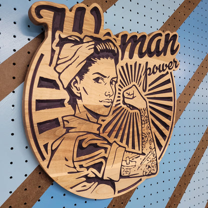 Angled shot of a custom hardwood Woman Power sign with updated Rosie the Riveter relief carving on a blue pegboard.