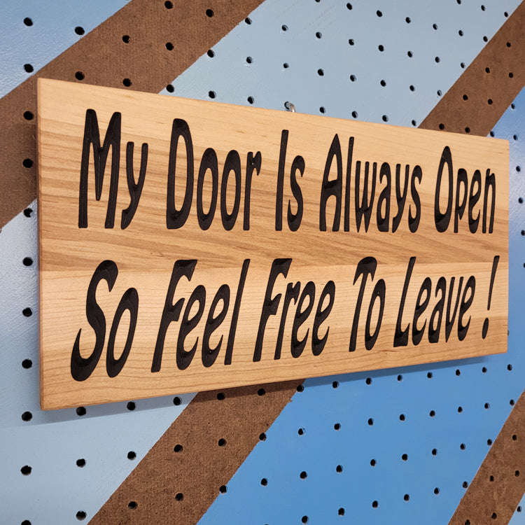 Angled view of a relief carved hardwood sign featuring the text: My Door Is Always Open So Feel Free To Leave!