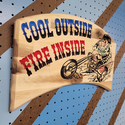 Angled shot of a relief carved hardwood sign with a retro monster chopper trike design and Cool Outside, Fire Inside text on a blue and brown striped pegboard.