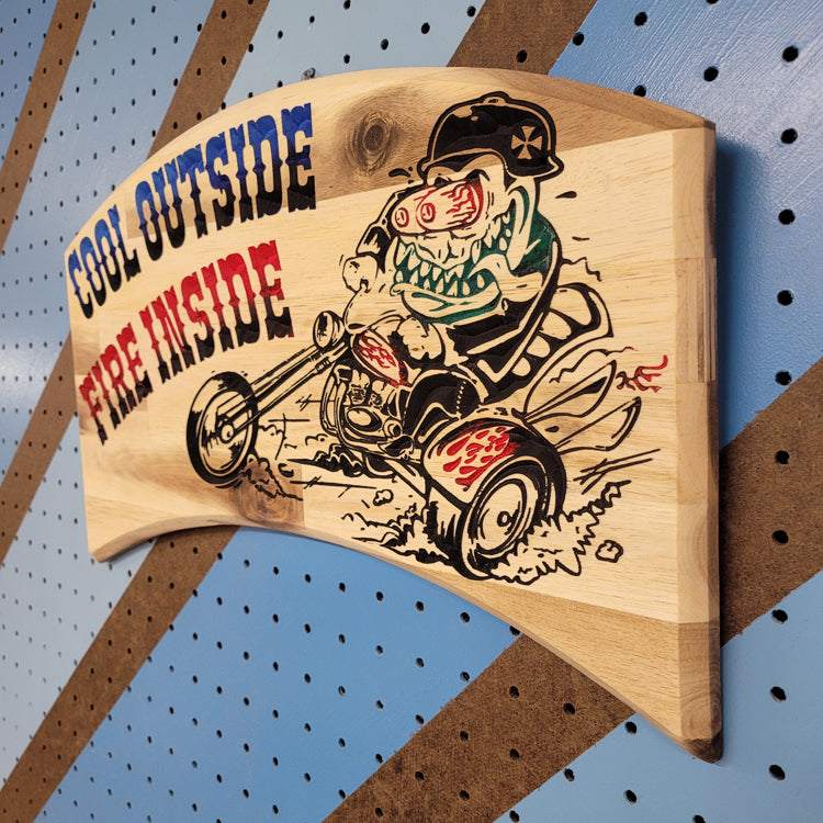 Angled shot of a relief carved hardwood sign with a retro monster chopper trike design and Cool Outside, Fire Inside text on a blue and brown striped pegboard.