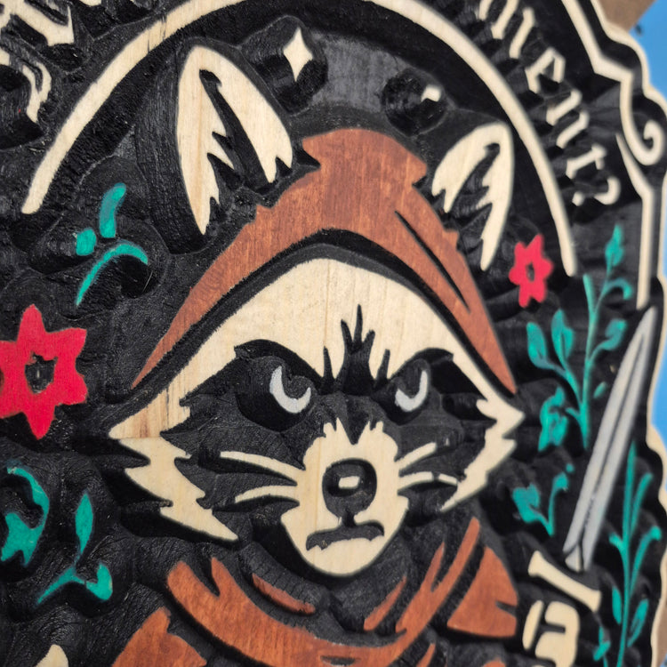 A close-up, detailed shot of a custom wood carving featuring a fantasy raccoon character. The image highlights the precise CNC-machined relief details of the raccoon's face, the texture of its hooded cloak, and the clean, deep grooves of the surrounding artwork.