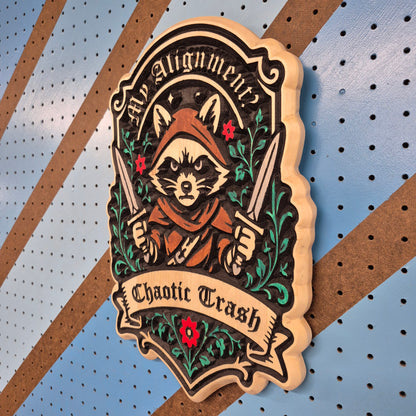 A solid hardwood carved sign featuring a fantasy rogue raccoon wearing a hooded cloak and holding twin daggers. The sign has a detailed circular frame that reads "My Alignment Is" with a banner below reading "Chaotic Trash," displayed against a blue and orange diagonal striped background.