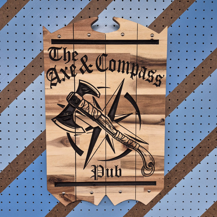 A vertical solid hardwood carved sign for "The Axe & Compass Pub". The detailed design features a battle axe layered over a compass rose and a frothy mug of ale, displayed against a blue and orange diagonal patterned background.