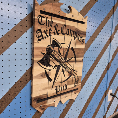 An angled, full-length shot of the Axe and Compass Pub wooden sign hanging on a wall, showcasing the clean CNC-machined relief cuts, dimensional depth, and natural grain of the hardwood.