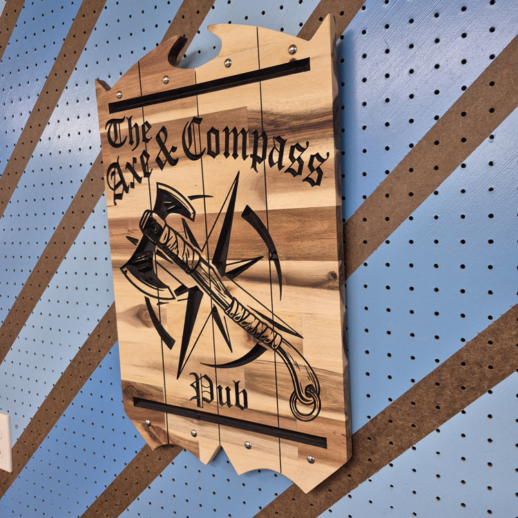 A side perspective view of the custom carved Axe & Compass tavern plaque, highlighting the precise edge work, the smooth finish of the wood, and how the carving catches the light.