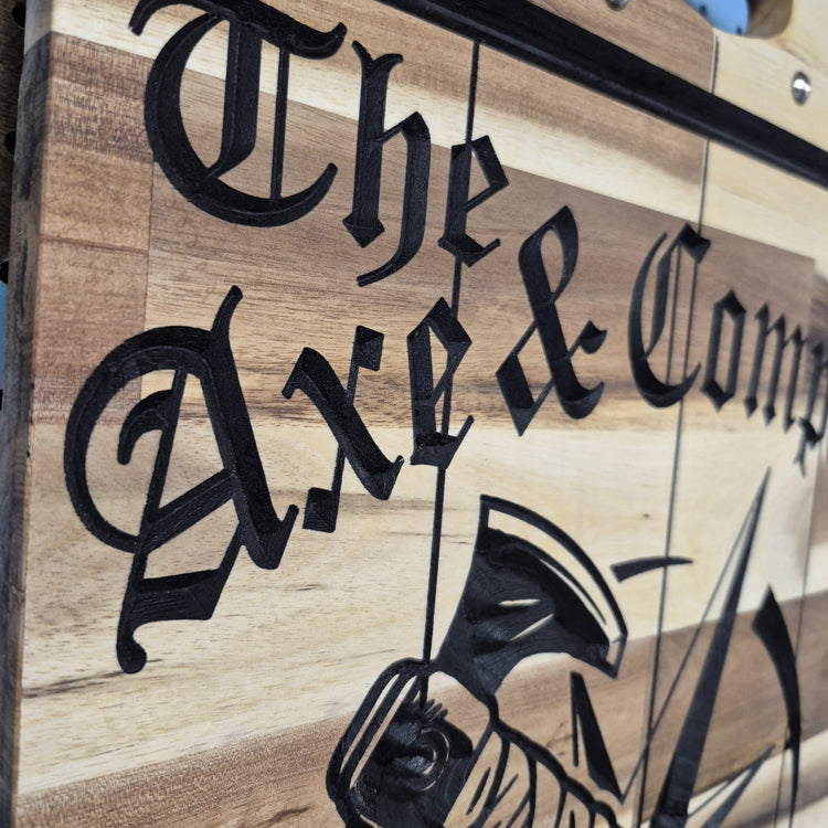 A tight close-up shot focusing on the top section of the sign, highlighting the deeply engraved text reading "The Axe & Compass" and the intricate, rustic wood grain textures within the carving.