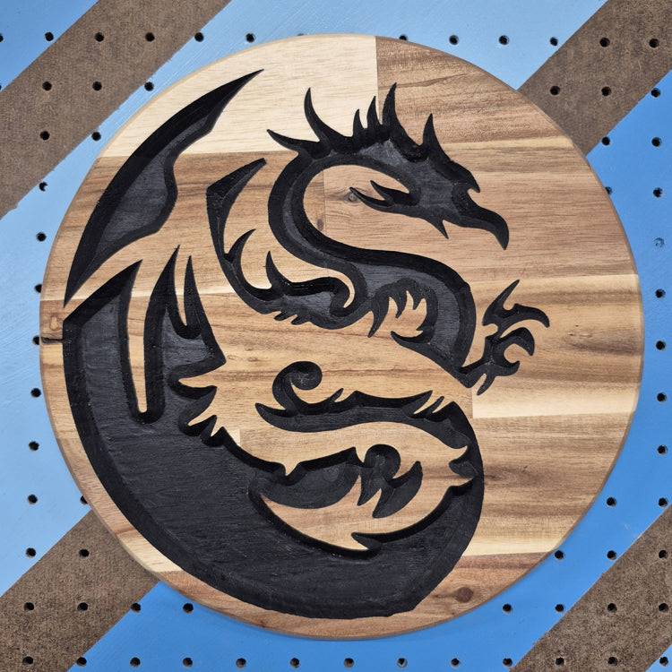 A circular solid hardwood carved sign featuring the "Yin Dragon" design. The carving depicts a stylized dragon forming a yin-yang symbol shape, with hand-painted black accents highlighting the grain, set against a blue and brown diagonal striped pegboard background.