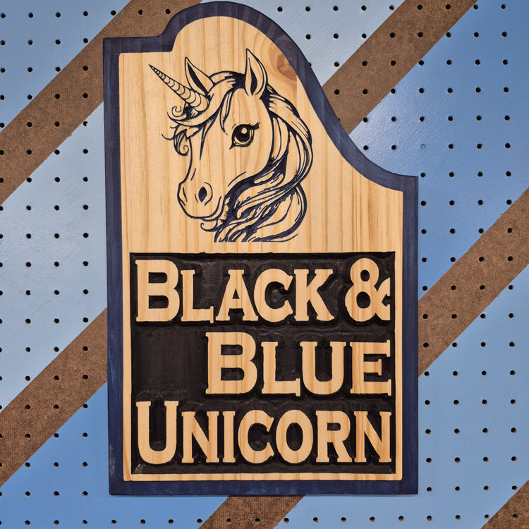 A vertical solid hardwood carved sign featuring a detailed fantasy unicorn head design. The precise CNC carving includes crisp hand-painted black and dark blue accents highlighting the unicorn's mane, face, and horn, displayed against a blue and brown diagonal striped pegboard background.