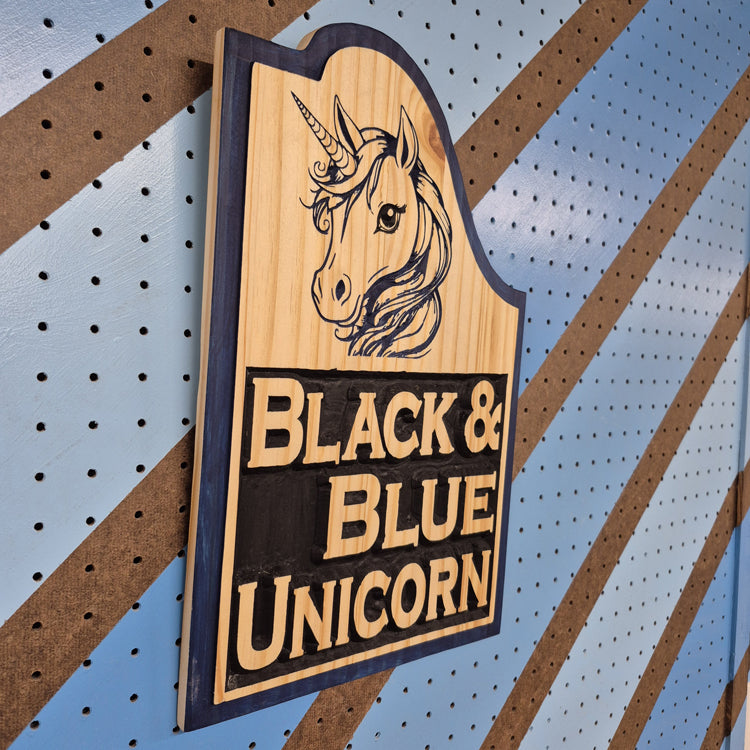 An angled side shot of the custom carved Black Blue Unicorn wooden plaque, showcasing the dimensional depth of the relief cuts and the striking contrast of the hand-painted accents against a striped pegboard wall.