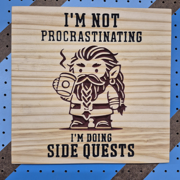 A square 12 by 12 inch solid hardwood carved sign featuring the word "Procrastinating" in a bold, stylized font. The precise CNC lettering features crisp hand-painted black accents, displayed against a blue and brown diagonal striped pegboard background.