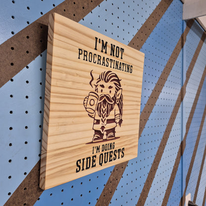 An angled side shot of the square Procrastinating wooden plaque, showcasing the deep dimensional relief of the CNC-machined cuts and the rich patterns of the natural hardwood grain against a striped pegboard wall.
