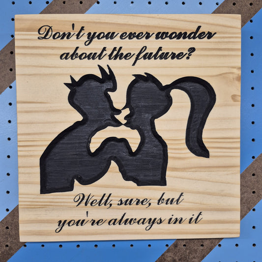 A square solid hardwood carved sign featuring the silhouettes of Fry and Leela from Futurama with the text "Don't you ever wonder about the future? Well, sure, but you're always in it." with hand-painted black accents against a blue and brown striped pegboard.