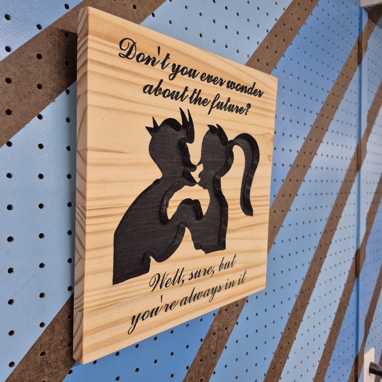 An angled side shot of the square sci-fi romance wooden plaque, showcasing the crisp depth of the CNC relief cuts and the rich patterns of the natural hardwood grain against a striped pegboard wall.