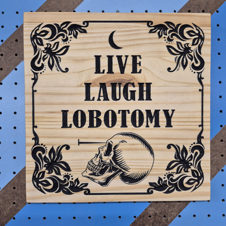 A square 12 by 12 inch solid hardwood carved sign featuring ornamental flourishes, an anatomical skull, and the text "LIVE LAUGH LOBOTOMY". The carving features hand-painted black accents against a blue and brown striped pegboard.