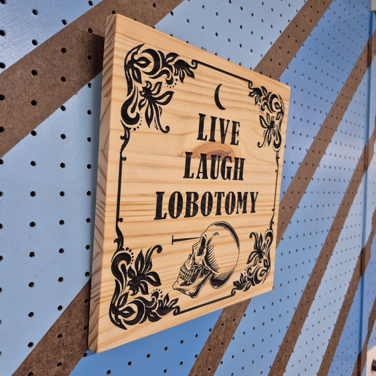 An angled side perspective of the square Live Laugh Lobotomy wooden plaque, showcasing the sharp depth of the CNC relief cuts and the rich patterns of the natural hardwood grain against a striped pegboard wall.
