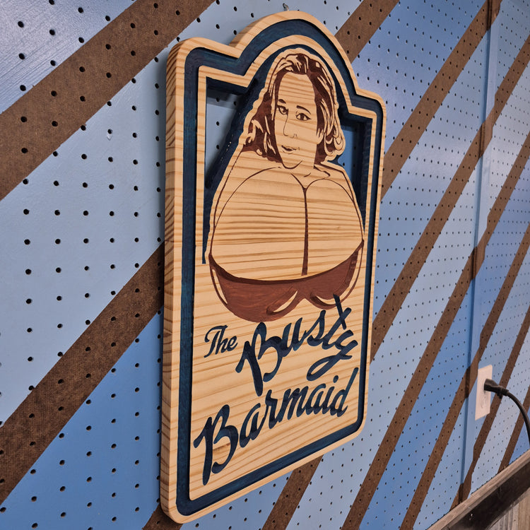 An angled side shot of the custom carved Busty Barmaid wooden plaque, showcasing the deep dimensional relief of the CNC cuts and the rich patterns of the natural hardwood grain against a striped pegboard.