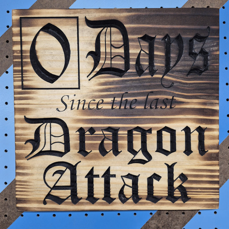 A square 12 by 12 inch solid hardwood carved novelty sign reading "0 Days Since the last Dragon Attack" in bold, rustic lettering. The carving features crisp hand-painted black accents against a blue and brown striped pegboard.