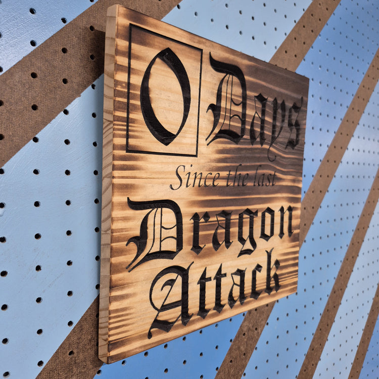 An angled side shot of the square Dragon Attack wooden plaque, highlighting the dimensional depth of the CNC relief cuts and the rich patterns of the natural hardwood grain against a striped pegboard wall.